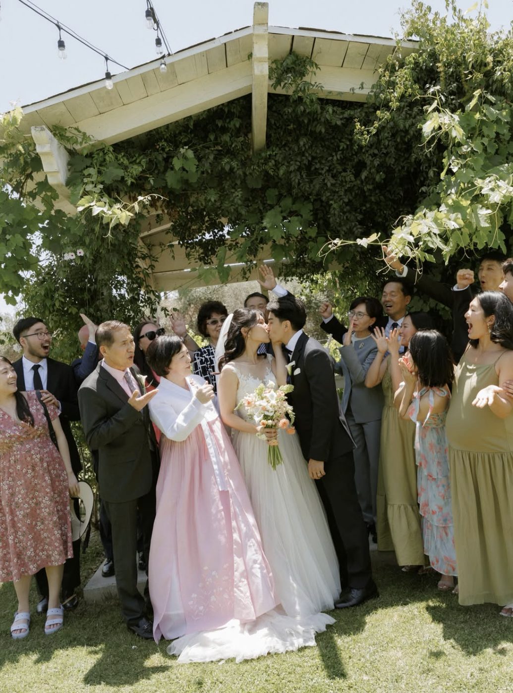 Family cheering as the couple kiss beneath a vine-covered arbor