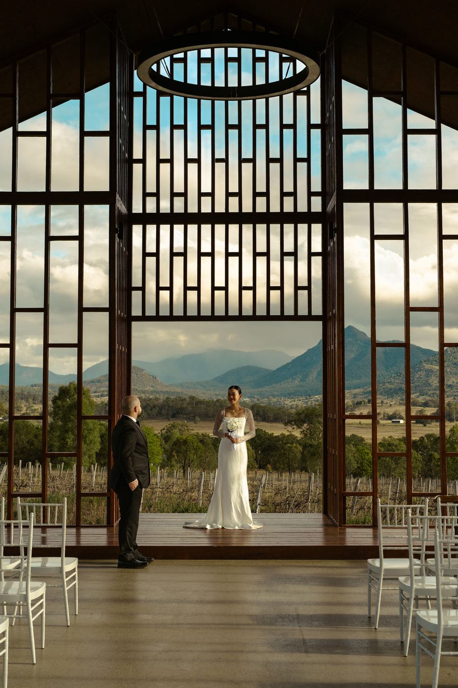 A couple facing each other at the altar of a glass chapel, mountains beyond