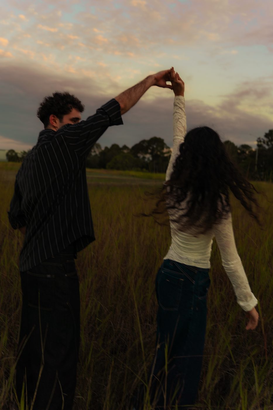 Two figures dancing in tall grass under pastel clouds at dusk