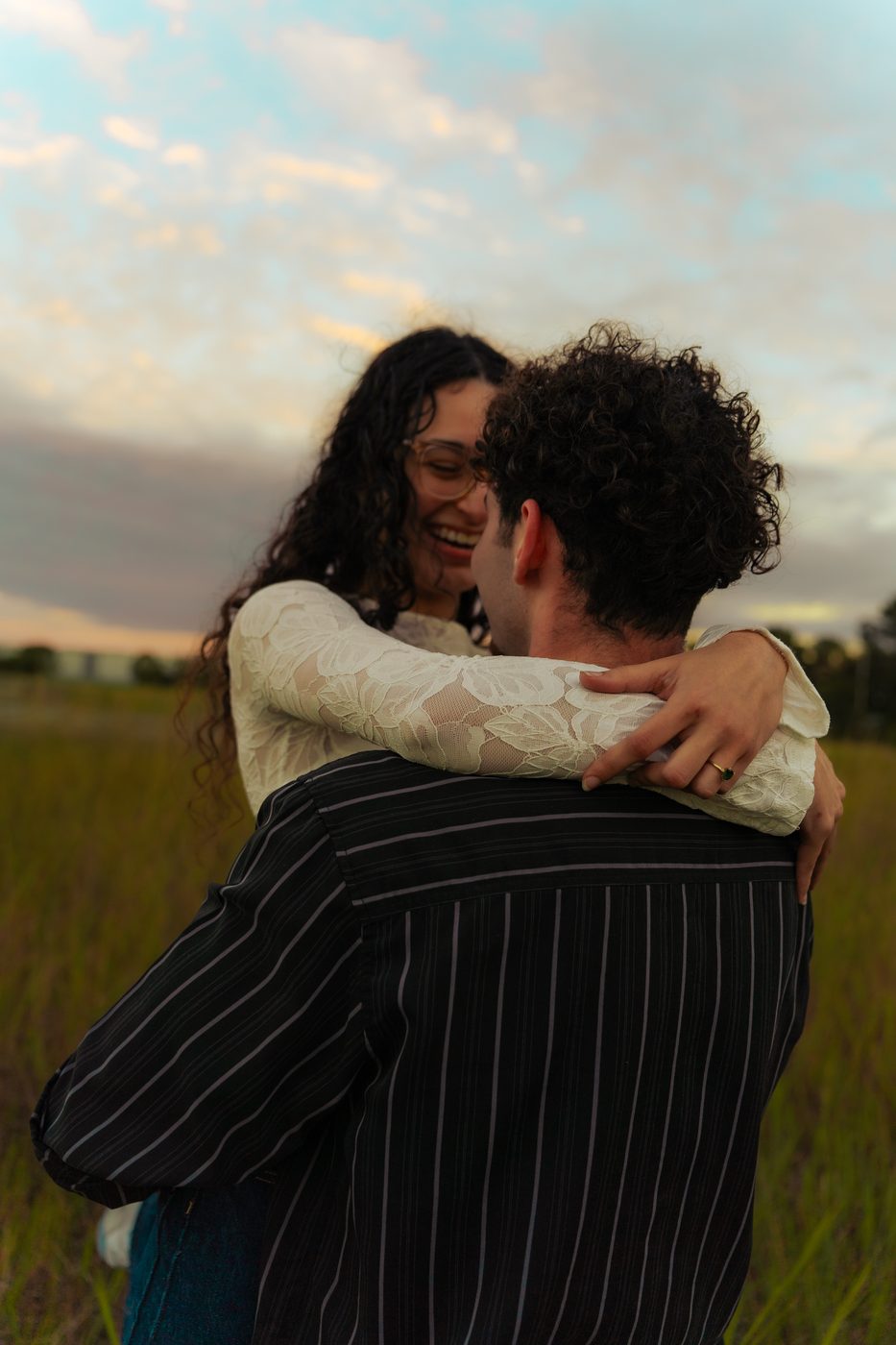 A couple holding each other in a field at sunset, she is laughing